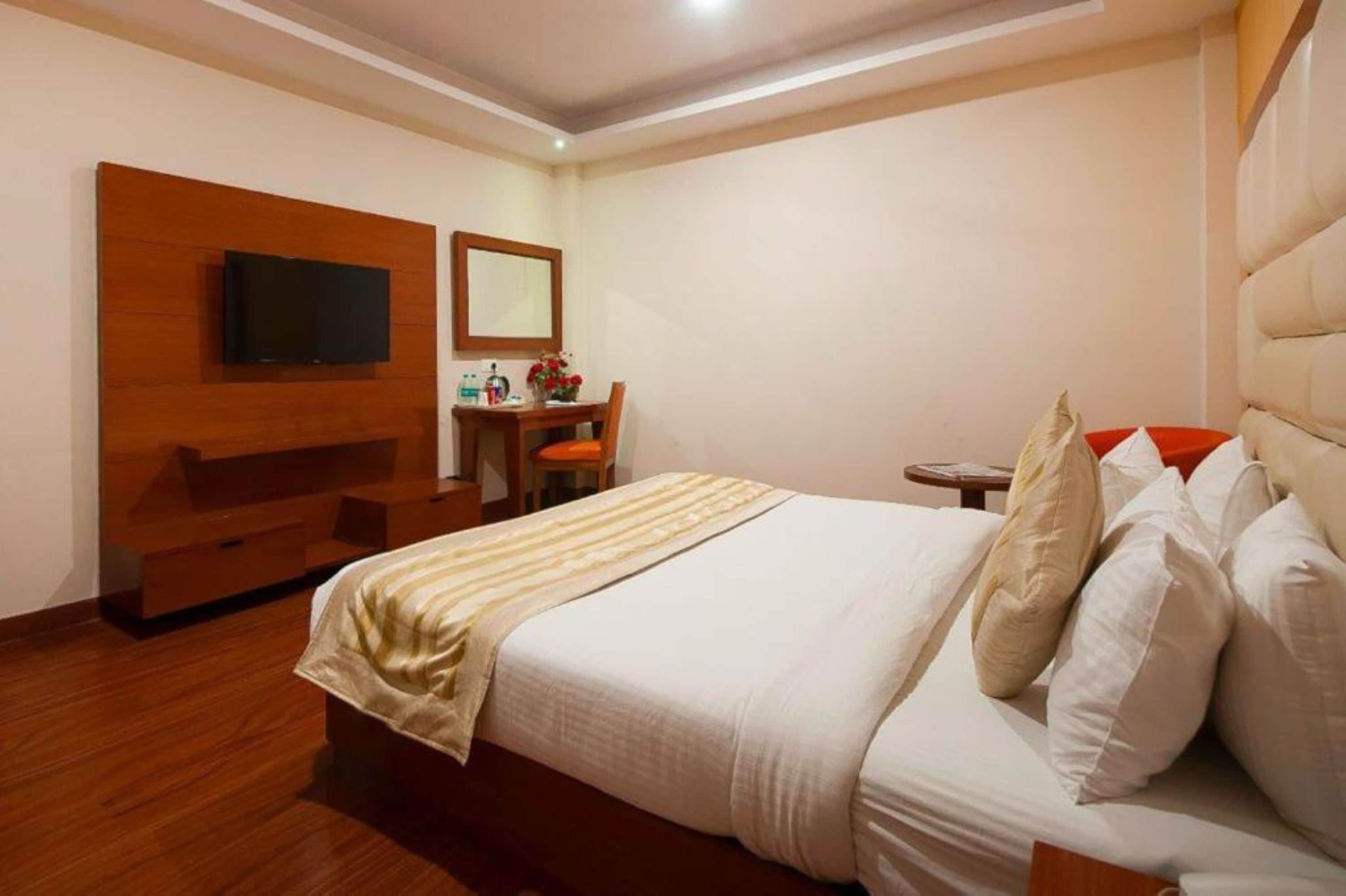 A hotel room with a bed, a TV mounted on a wooden panel, a desk with a chair, and a small table with a vase of flowers.