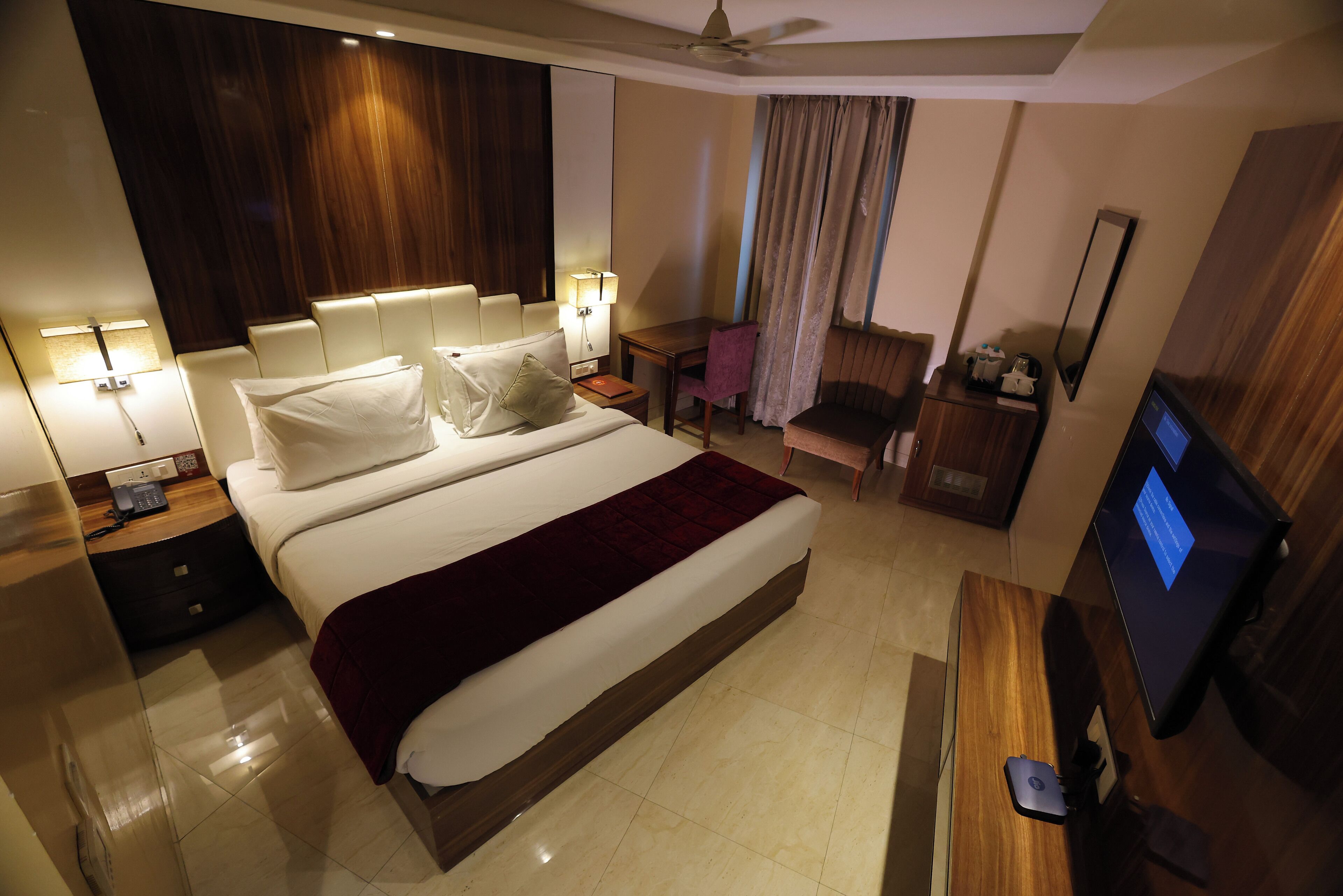 A hotel room with a bed, bedside tables, a desk with a computer, a chair, a TV, and a ceiling fan.