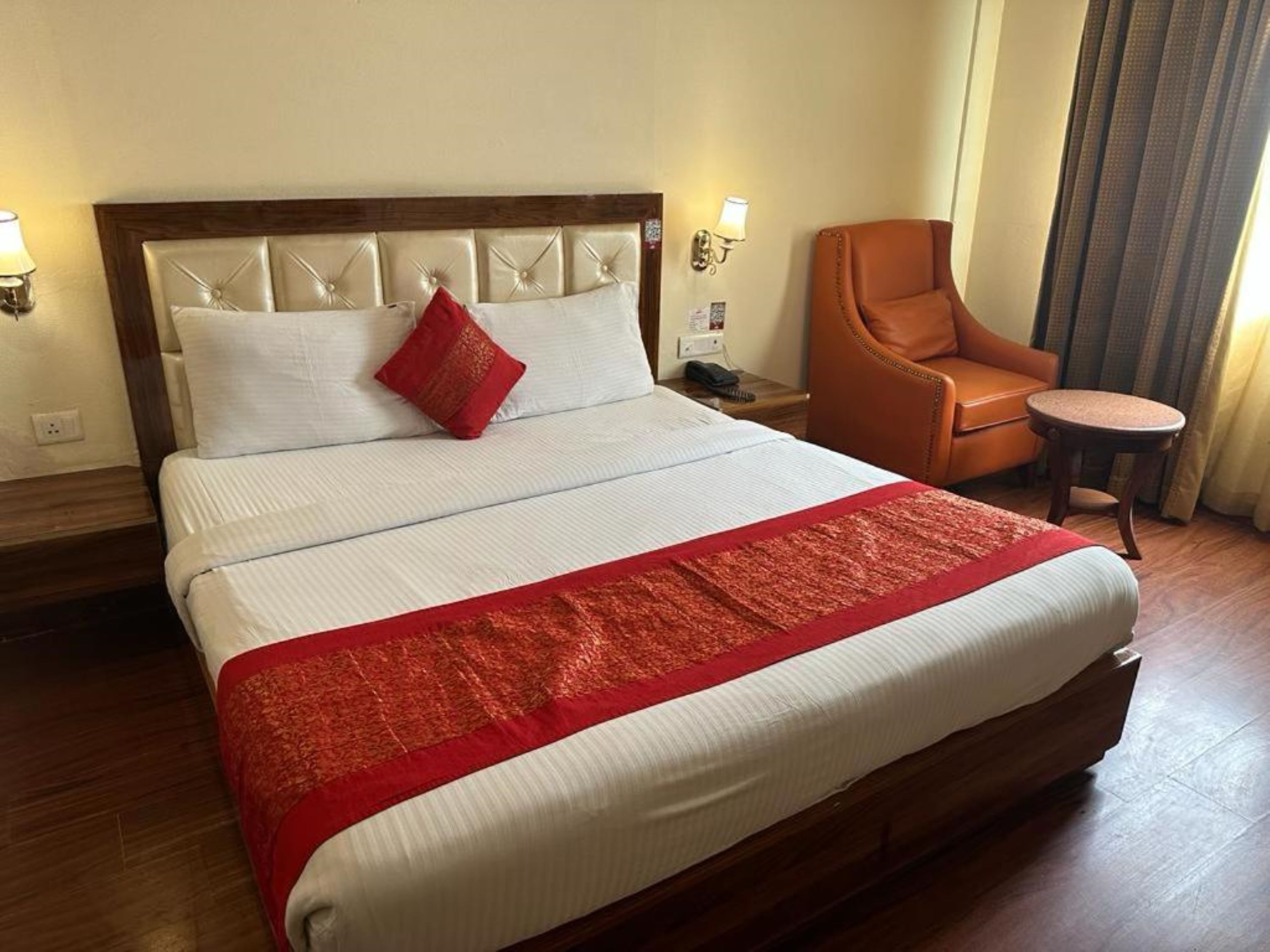 Ein Hotelzimmer mit einem großen Bett, einer roten Tagesdecke, einem Holzkopfende, einem braunen Ledersessel, einem kleinen runden Tisch und einem Nachttisch mit Lampe.