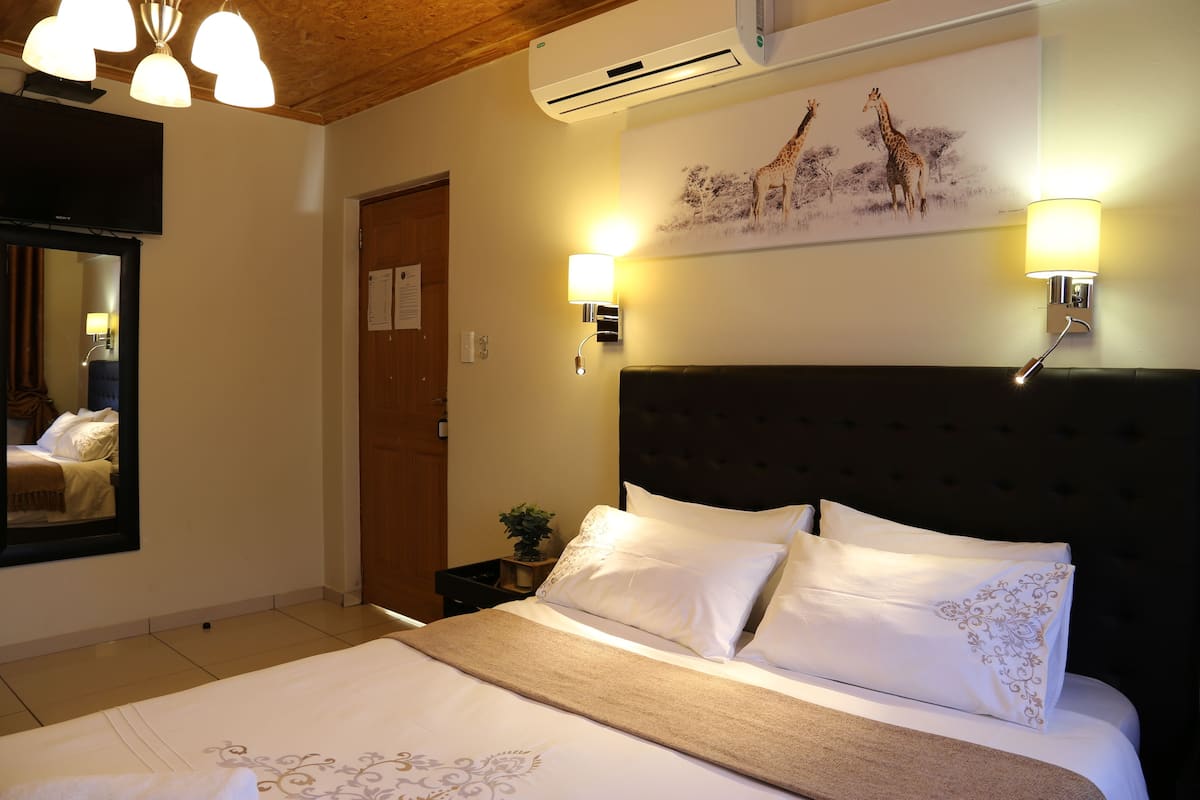 Habitación de hotel con una cama grande, cabecera, dos lámparas de pared, un equipo de aire acondicionado, un cuadro de jirafas y techo de madera.