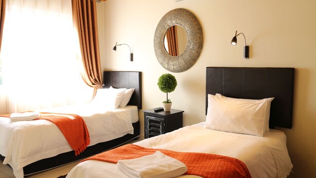 Quarto de hotel com duas camas, um espelho redondo, uma pequena planta em vaso e uma mesa de cabeceira com abajur.
