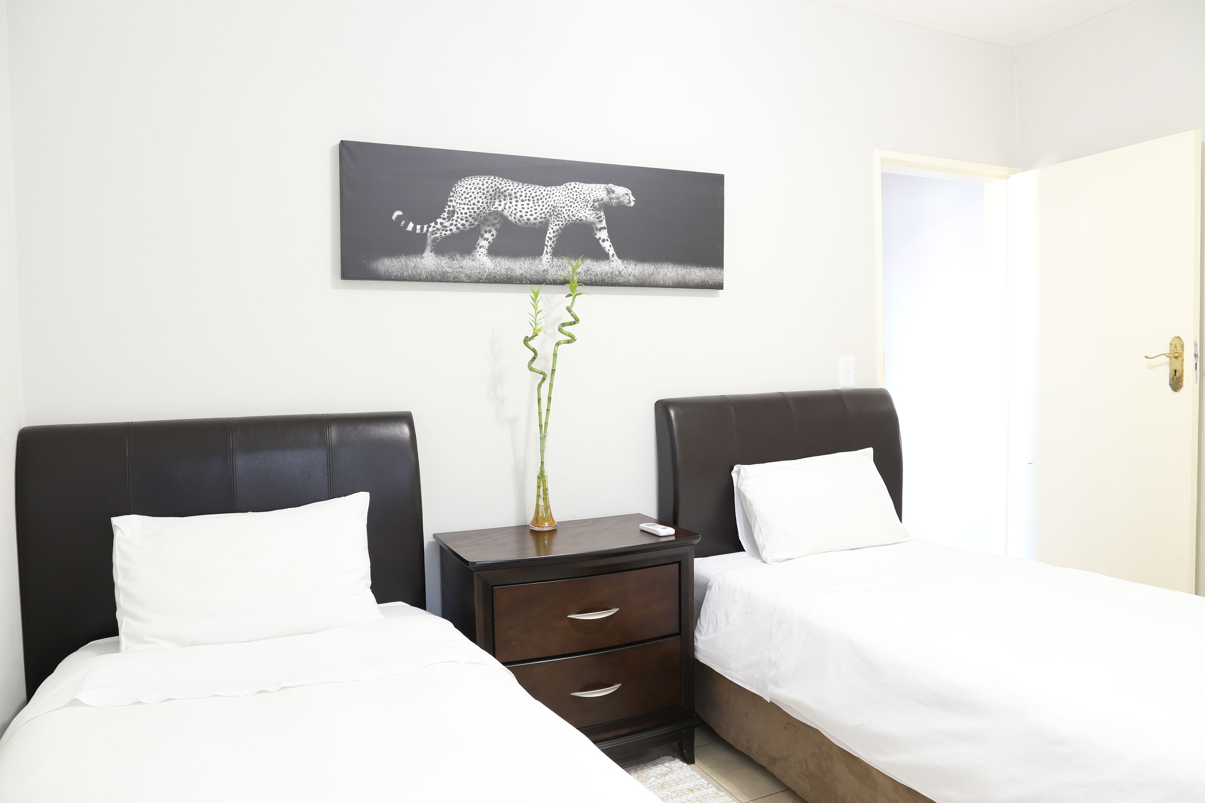 Habitación de hotel con dos camas, una mesita de noche con cajones, un florero con planta y un cuadro colgado en la pared con la imagen de un guepardo.