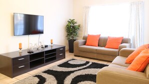 Uma sala de estar moderna com televisão de tela plana, painel de TV em madeira escura, sofá bege com almofadas laranja e tapete com padrão preto e branco.