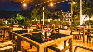 Un área de comedor al aire libre con mesas y sillas de mimbre, rodeada de follaje y un edificio al fondo.