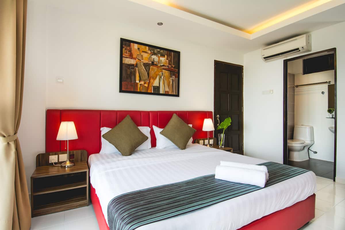 A hotel room with a large bed, red headboard, two bedside tables with lamps, a painting on the wall, and an air conditioning unit.