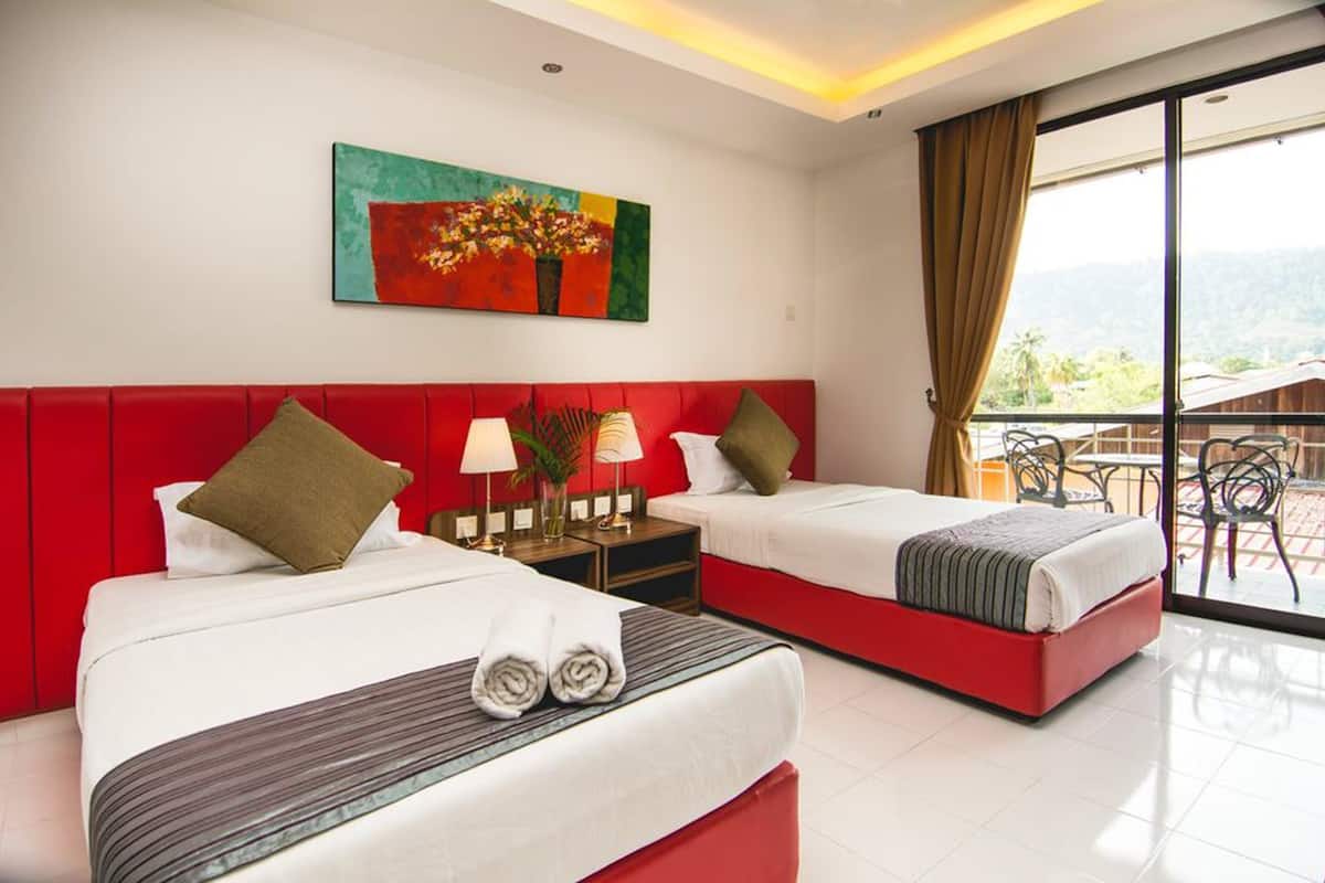 A hotel room with two beds, a red headboard, a painting, a nightstand with lamps, and a balcony with chairs.