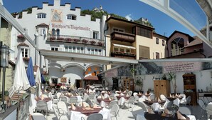 An outdoor café with tables and chairs, a building with a sign reading "Walther von Vogelweide," and a view of a mountain. - Walther von der Vogelweide (Chiusa)