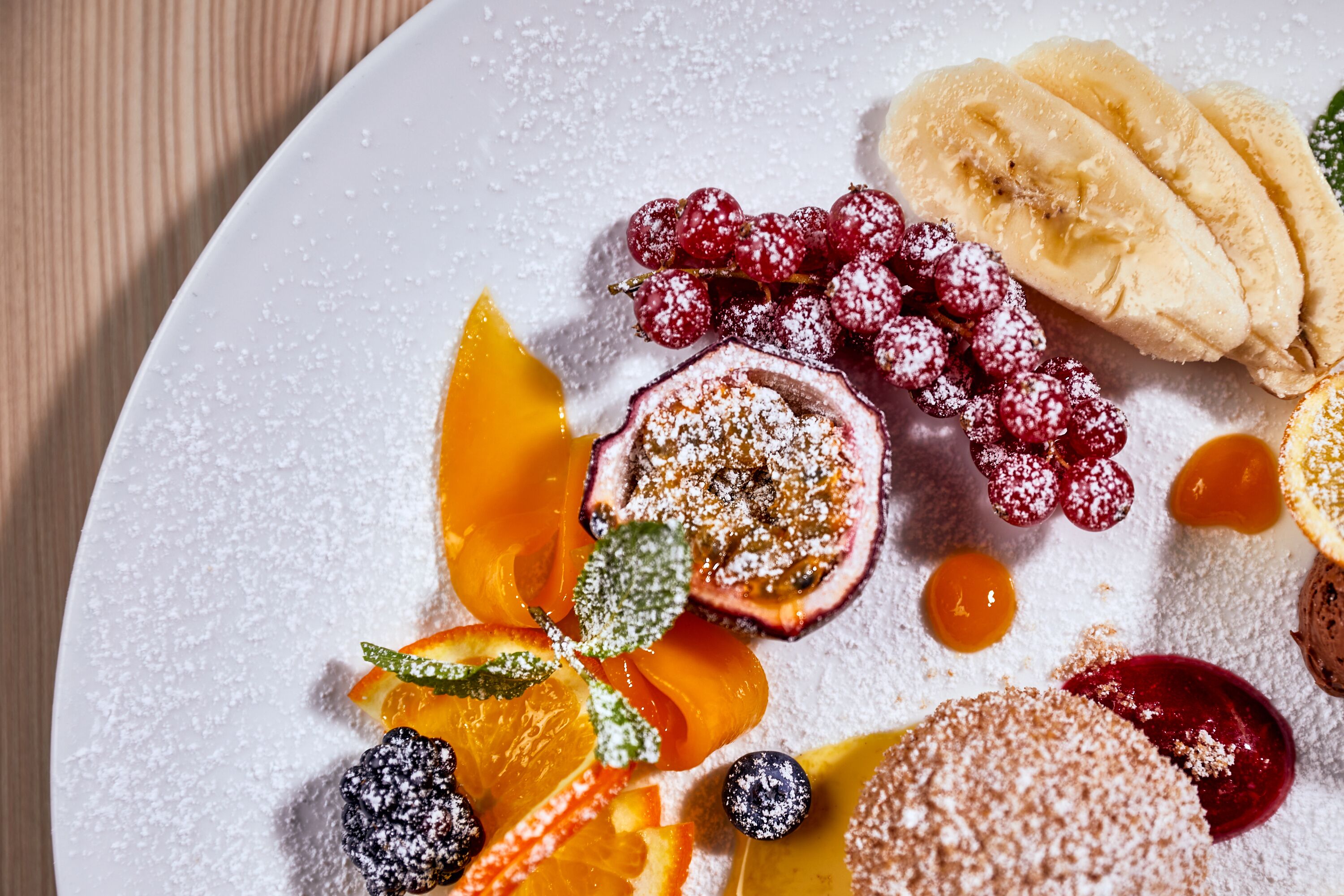 Un plato de postre con rodajas de plátano, fresas, arándanos y media fruta, posiblemente un melocotón o nectarina, espolvoreado con azúcar glas.
