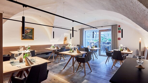 A modern dining area with wooden tables and chairs, a large window with a view of the outdoors, and a painting on the wall. - Walther von der Vogelweide (Chiusa)