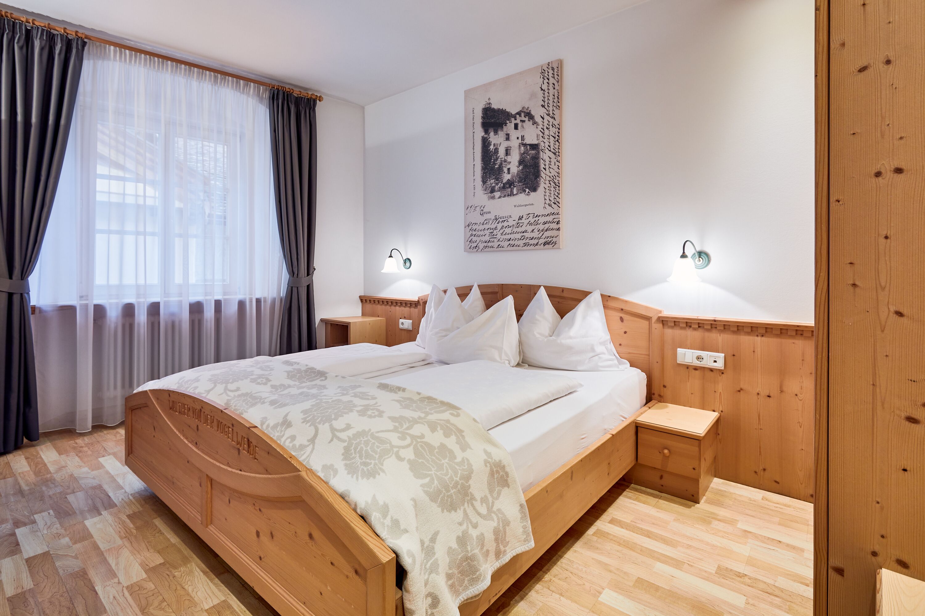 Een houten bed met hoofdbord, twee nachtkastjes, een wandlamp en een raam met gordijnen.