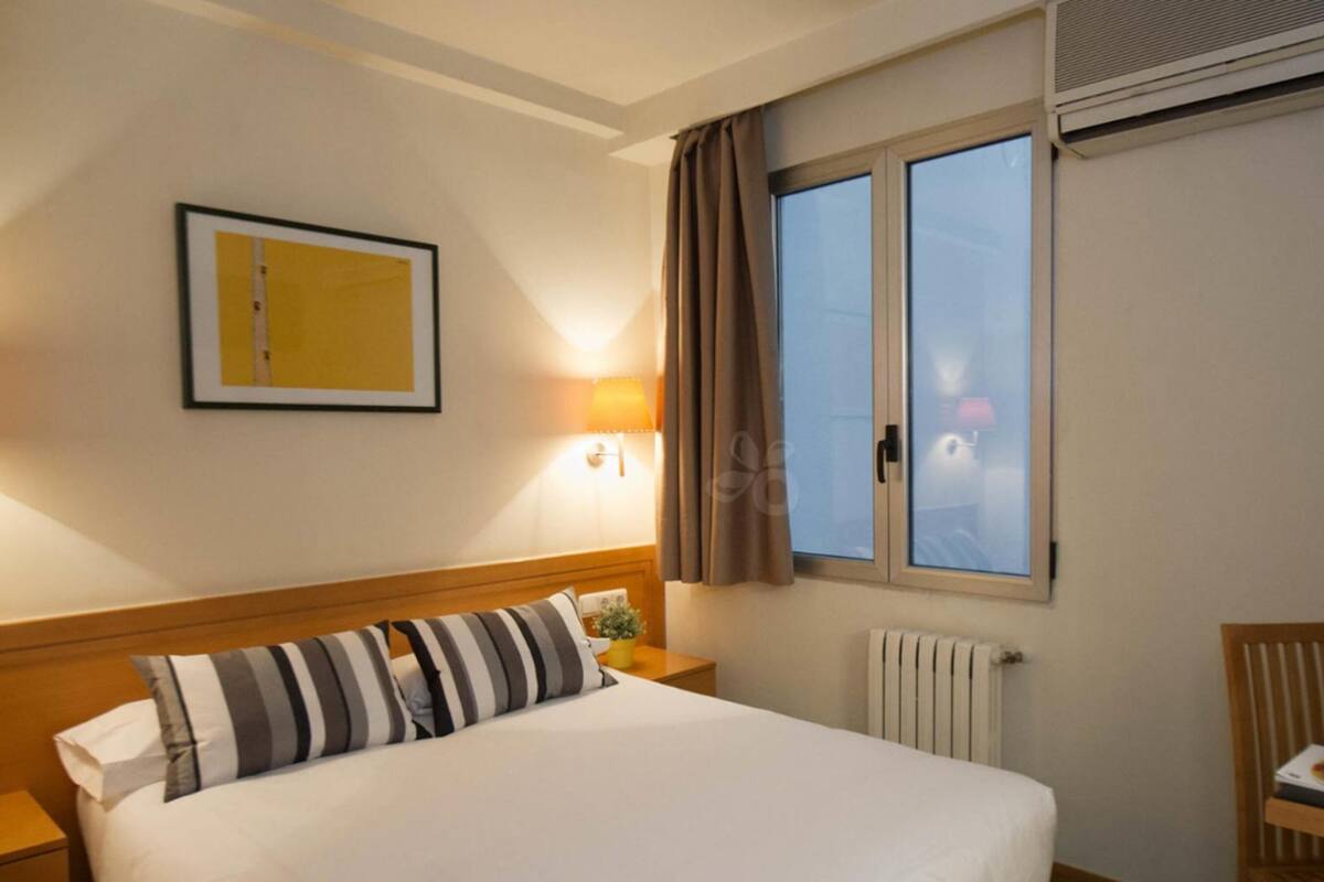 Een hotelkamer met een bed, nachtkastje, lamp, raam met gordijnen, verwarming en een lijst aan de muur.