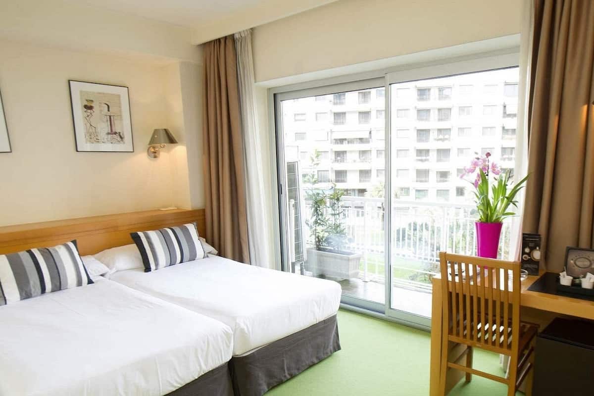 Een hotelkamer met twee bedden, een bureau met stoel, een raam met uitzicht op gebouwen en een plant in een pot op de vensterbank.