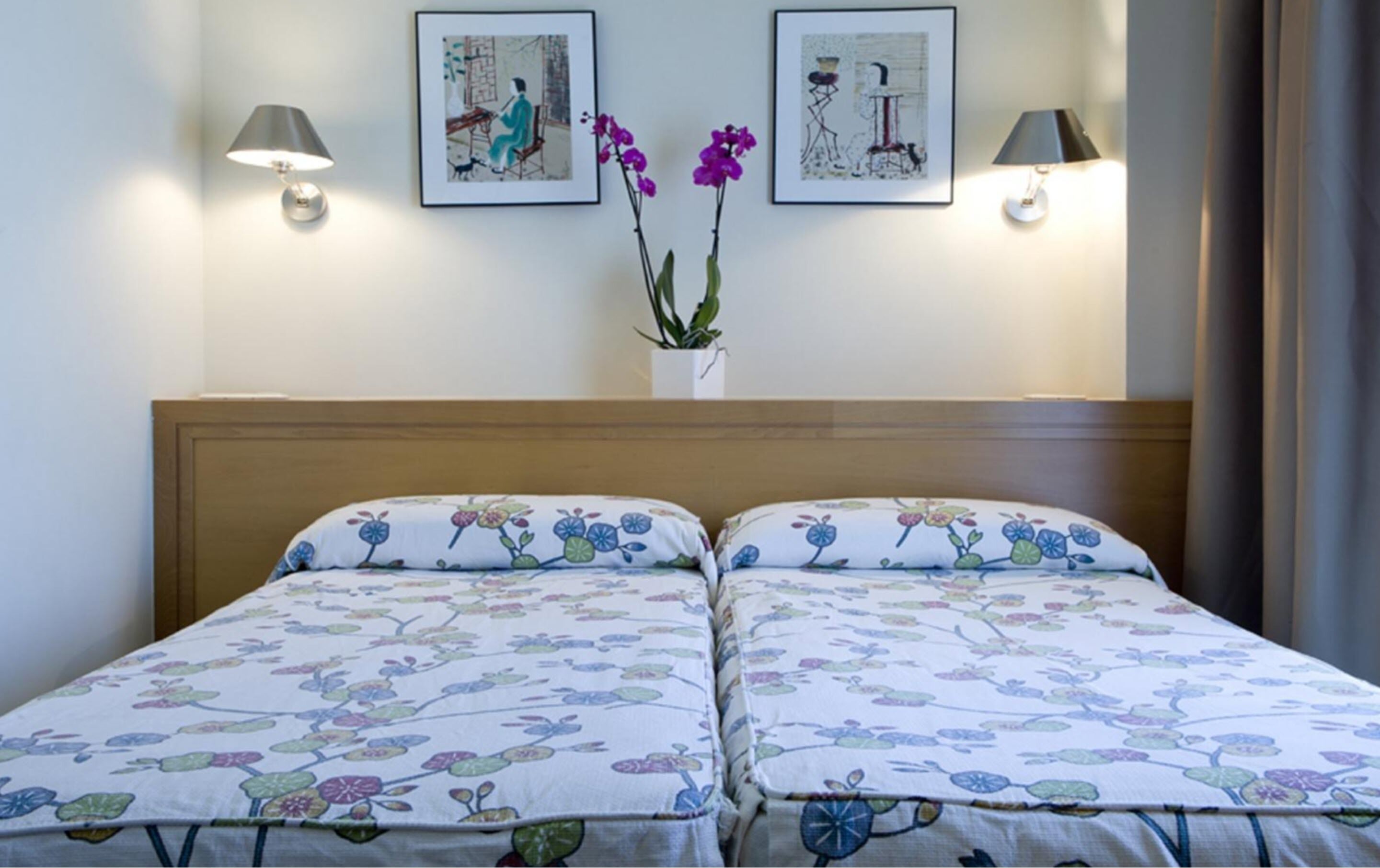 A double bed with floral bedding, two wall-mounted lamps, and two framed artworks above it.