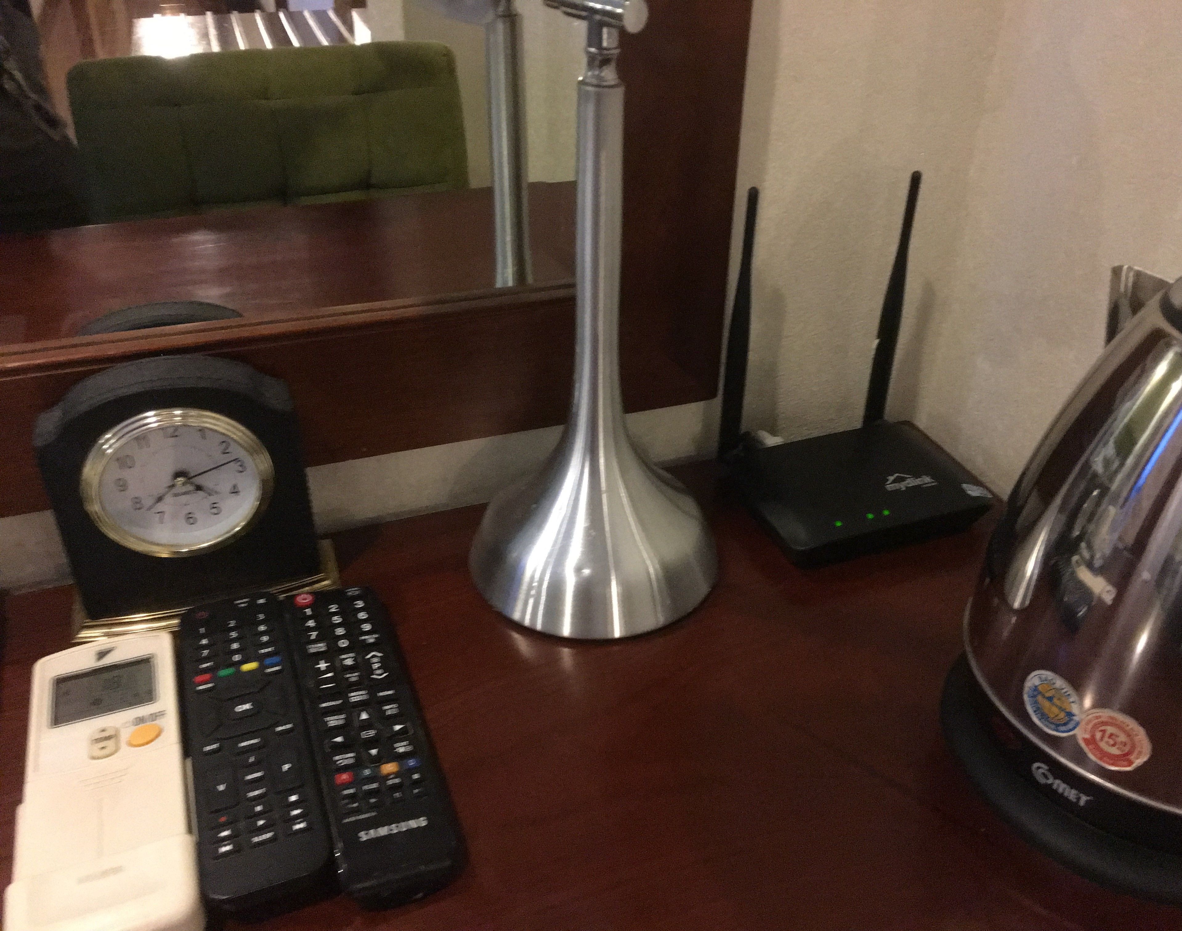 Un escritorio de madera con un reloj, un control remoto de Samsung, un enrutador negro y una lámpara de metal.