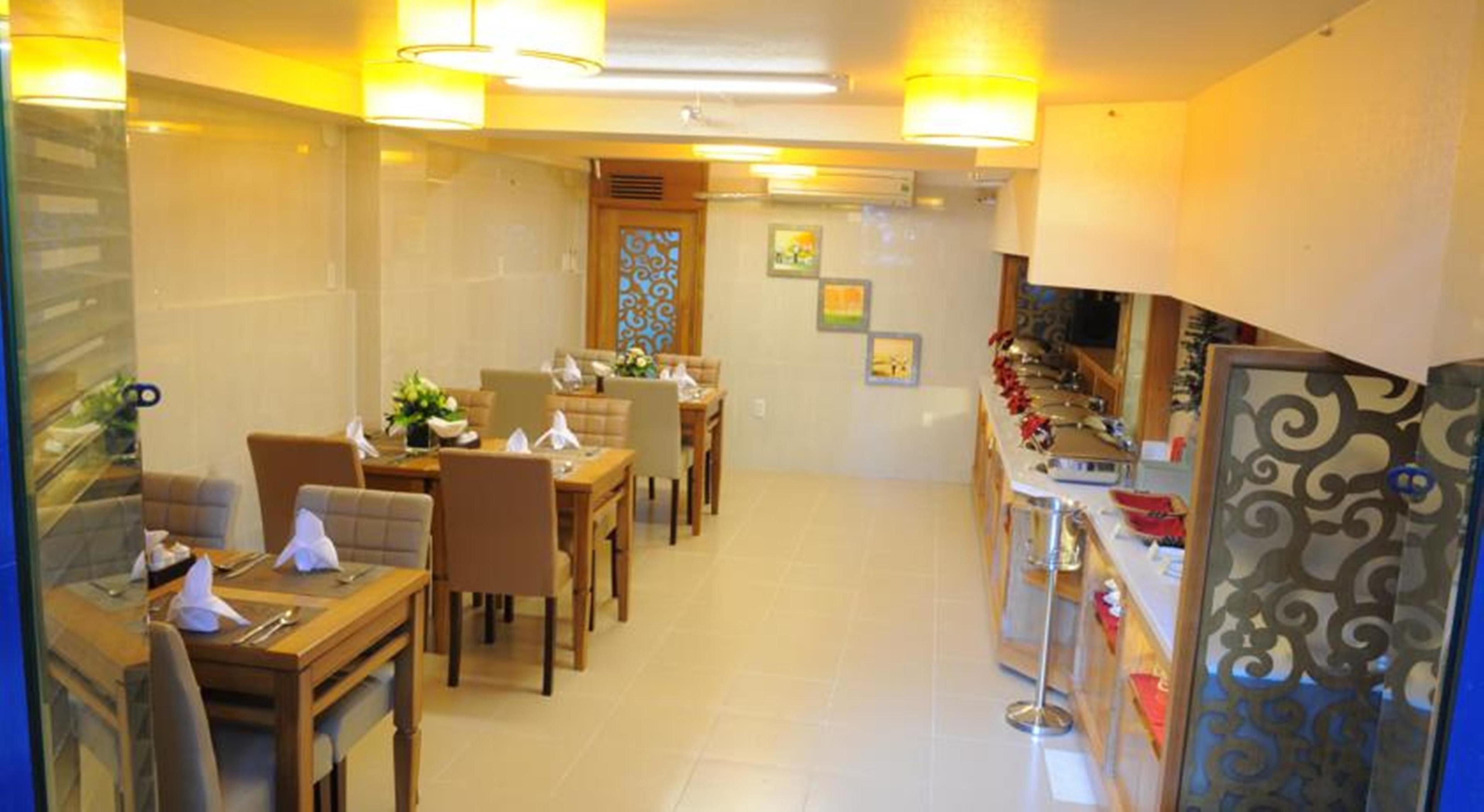 Une salle à manger d’hôtel avec plusieurs tables et chaises, un coin cuisine doté d’un comptoir et d’ustensiles, et un mur décoratif orné de tableaux encadrés.