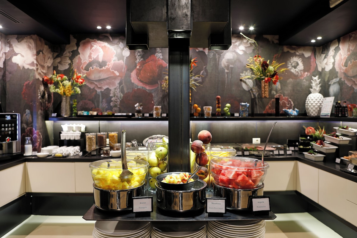 Frokostbuffet med frukt i glassboller foran en vegg med store blomstermotiver.