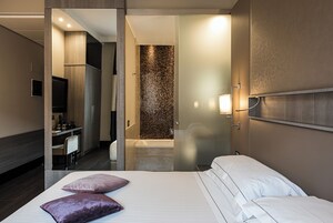 A modern hotel room with a large bed, a desk with a chair, a television, and a bathroom with a tiled wall.