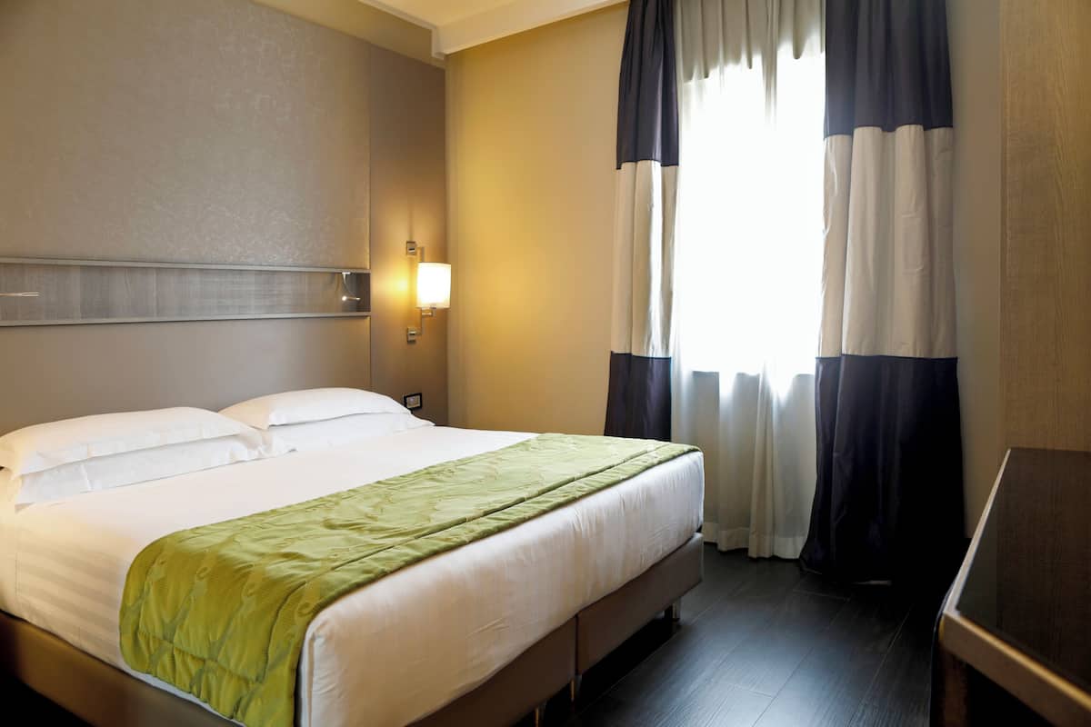 Ein Hotelzimmer mit Bett, Nachttisch, Lampe und einem Fenster mit Vorhängen.