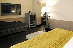 A hotel room with a bed, a sofa, a TV, a lamp, and a small table with a lamp.