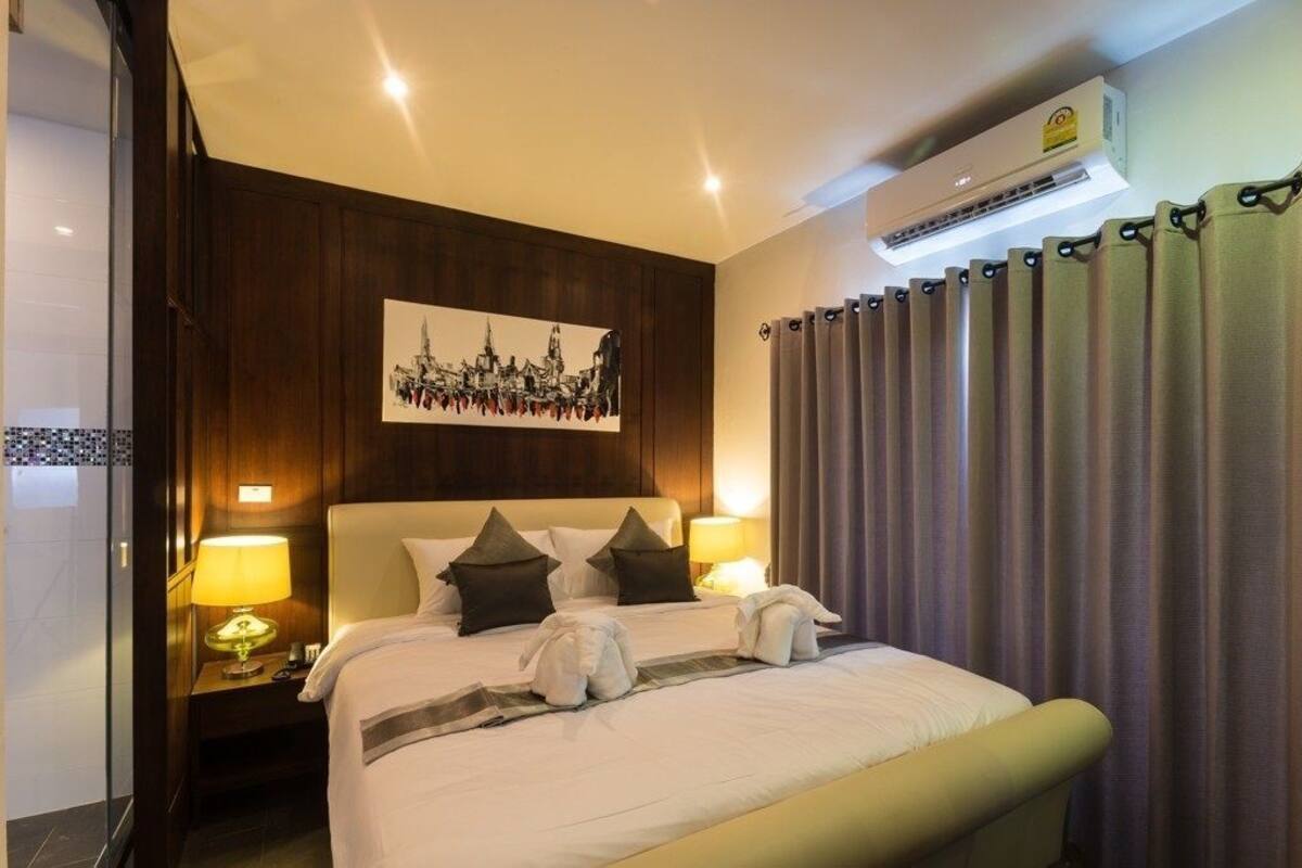 A hotel room with a bed, a headboard, a nightstand with a lamp, a wall-mounted air conditioner, and a curtain.