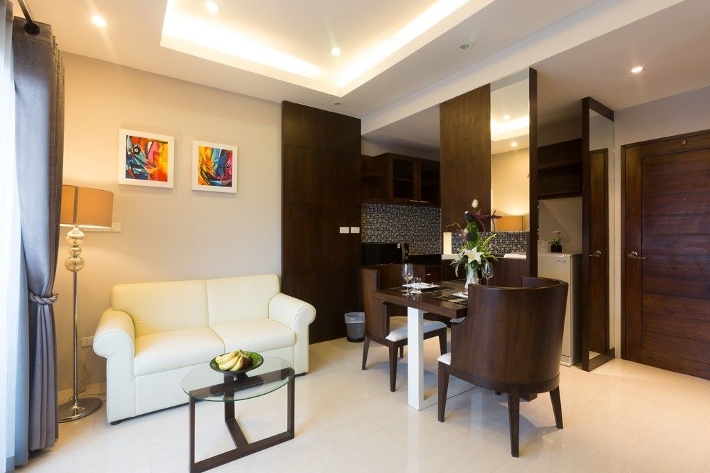 A modern living room with a white sofa, a glass coffee table, and a dining area with a dark wooden table and chairs. There are two abstract paintings on the wall, a floor lamp, and a kitchen visible through an open door.