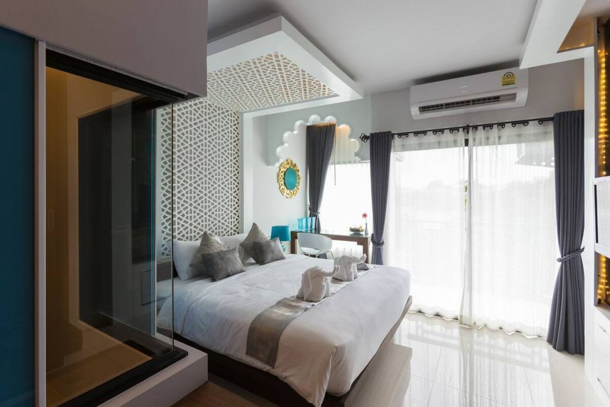 A modern hotel room with a large bed, a glass partition, a decorative wall, a ceiling light, and a view of the outdoors.