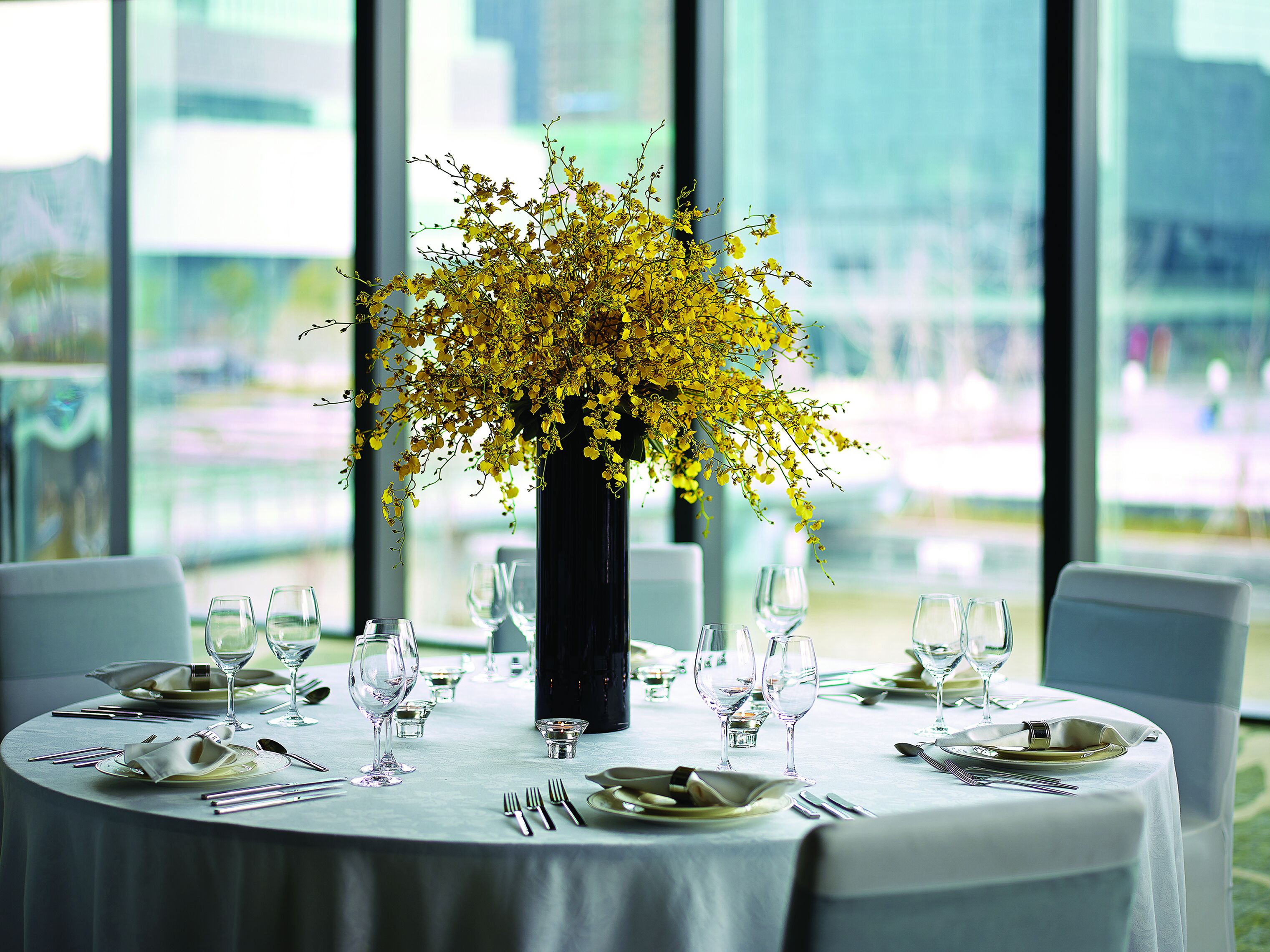 Una mesa redonda para comer con mantel blanco, servida con platos, cubiertos y copas de vino. Un jarrón negro con flores amarillas está colocado en el centro de la mesa.