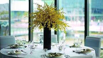 Een ronde eettafel met een witte tafeldoek, gedekt met borden, bestek en wijnglazen. In het midden van de tafel staat een zwarte vaas met gele bloemen.