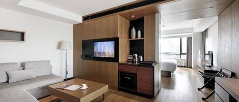 A modern hotel room with a sofa, coffee table, TV, kitchenette, and a view of the city.