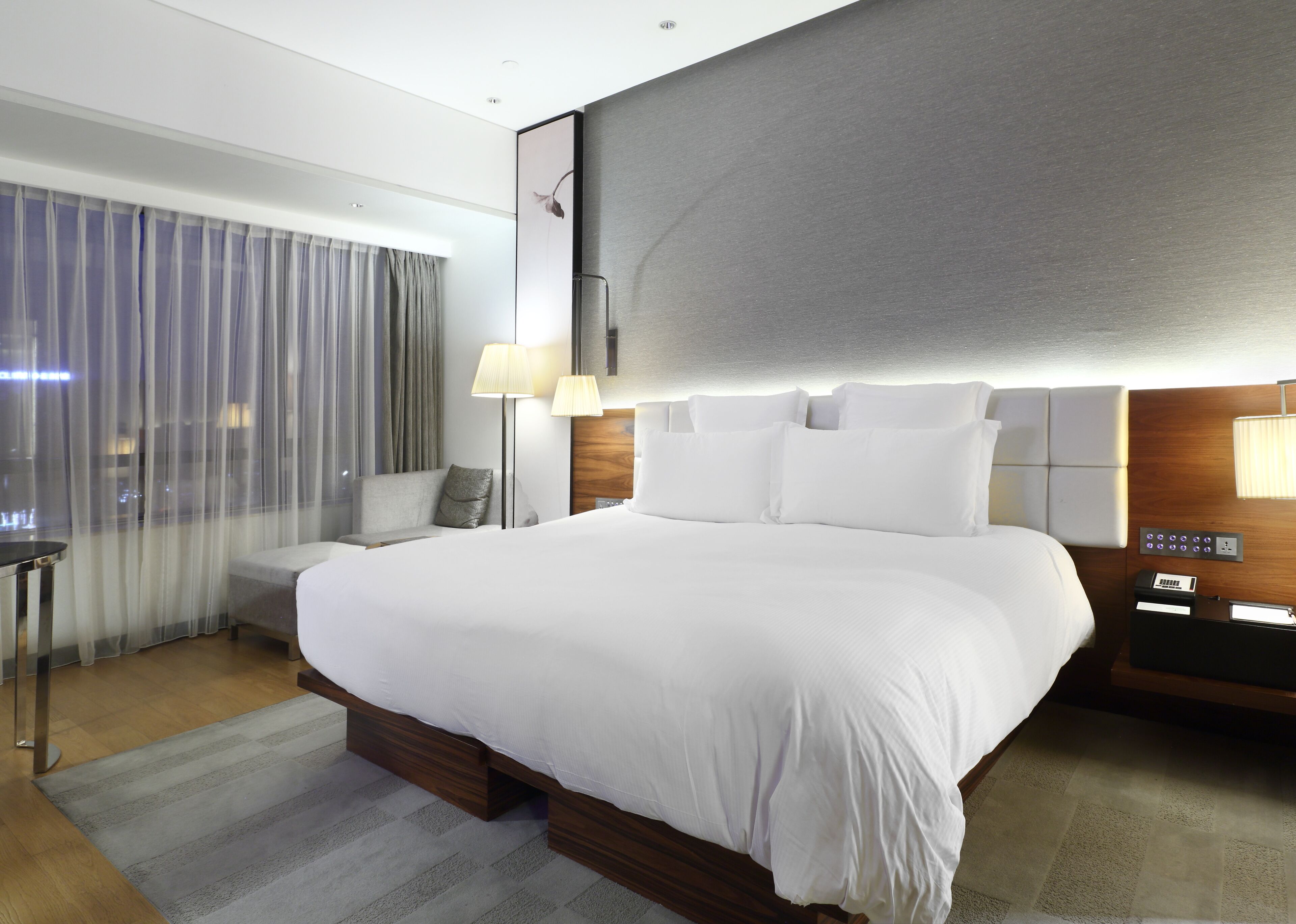 A modern hotel room with a large bed, a bedside table, a lamp, and a view of the city through the window.