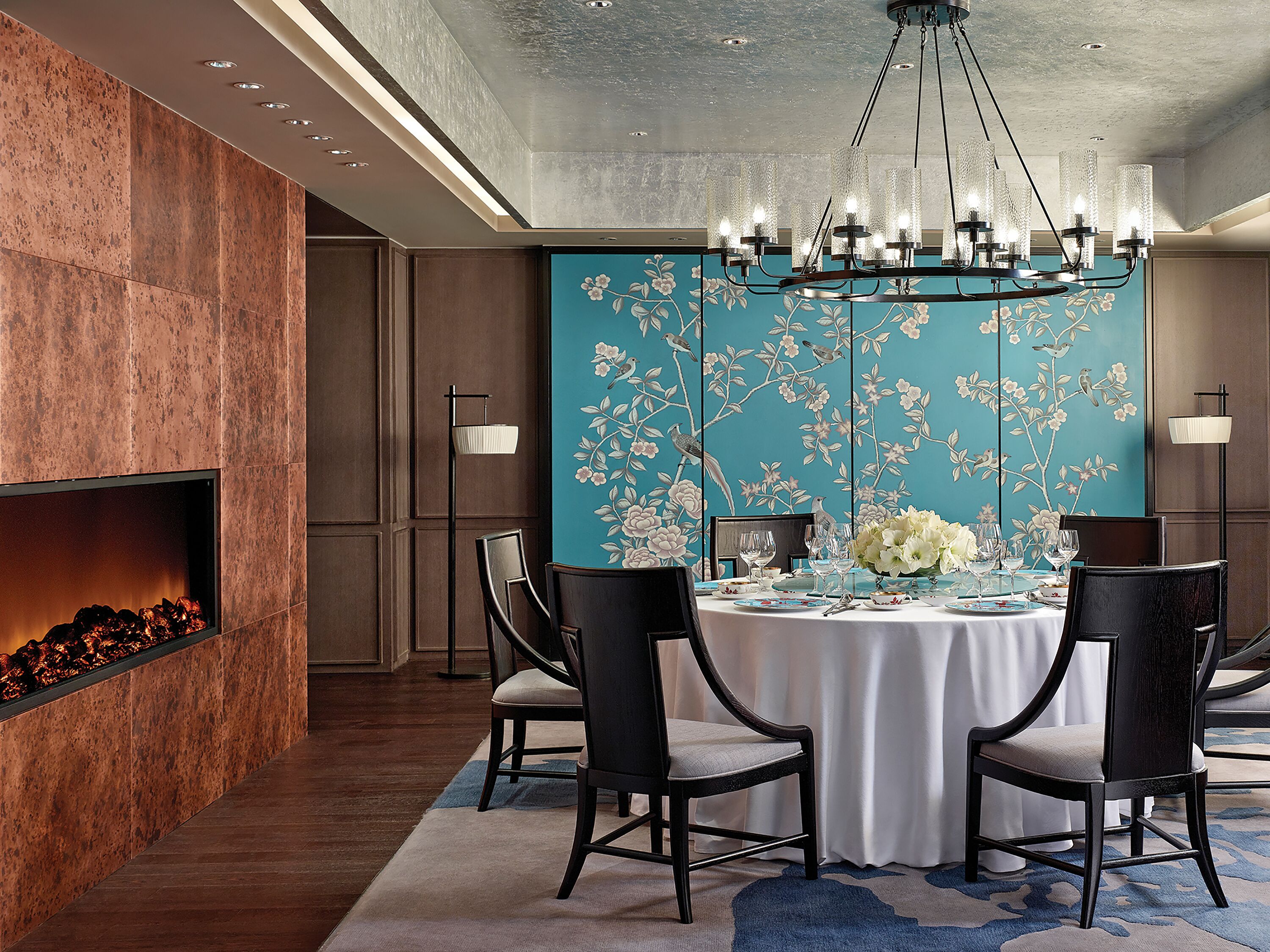 A dining area with a round table set for four, surrounded by dark chairs. A fireplace is visible on the left, and a decorative wall with floral patterns is behind the table.