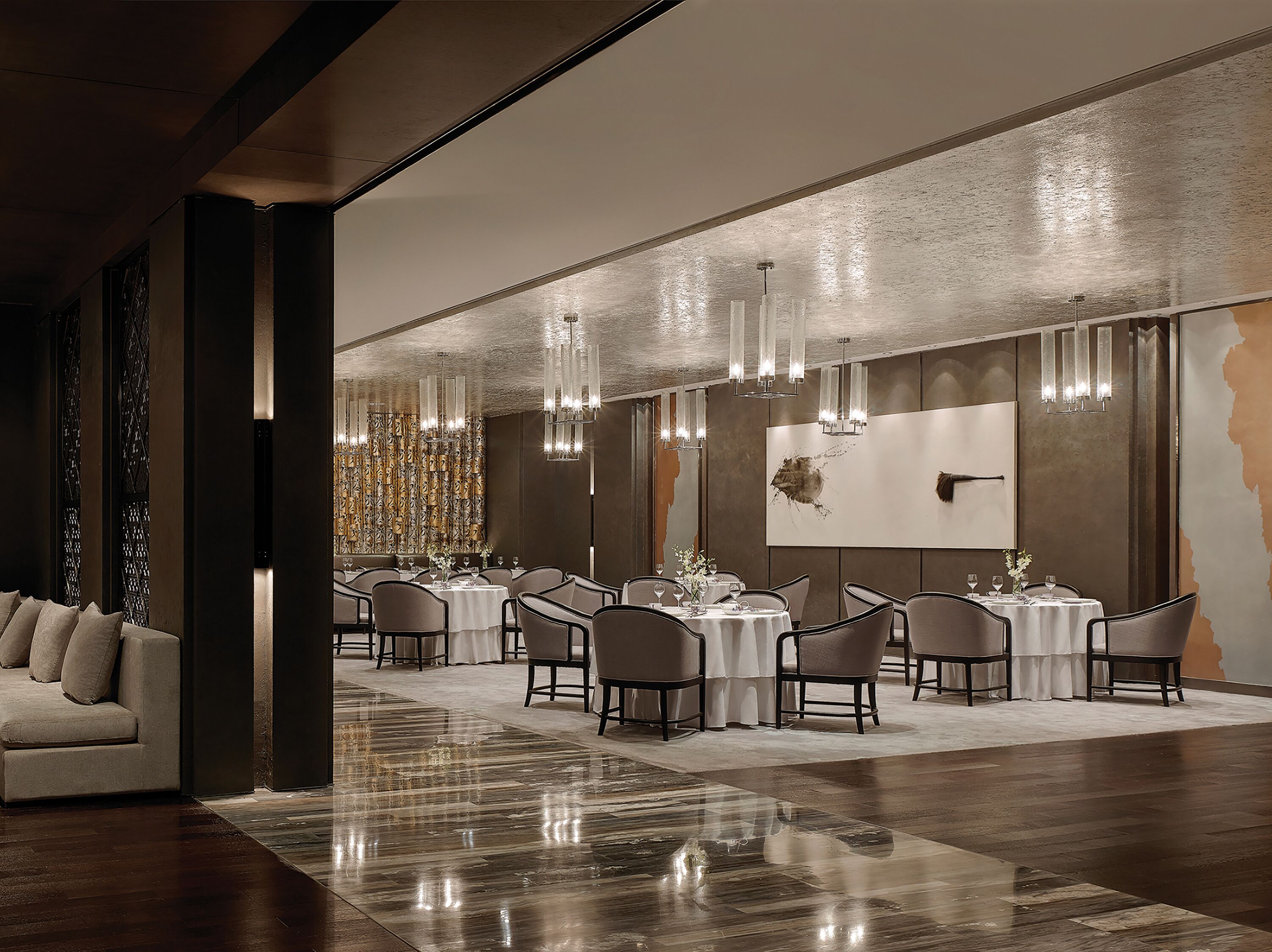 A modern dining area with multiple tables and chairs, elegant chandeliers, and a large abstract painting on the wall.