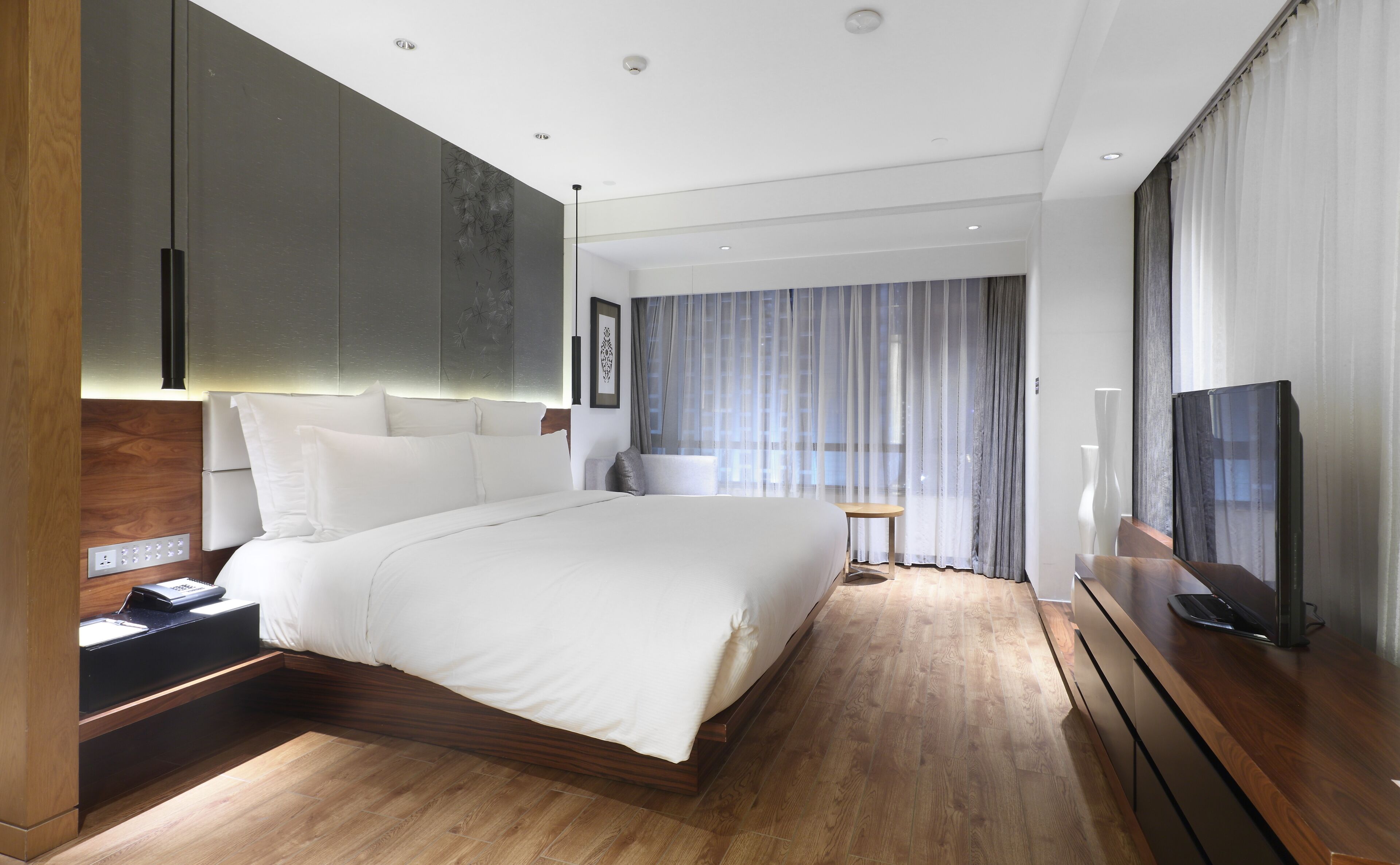 A modern hotel room with a large bed, wooden flooring, a desk with a computer, a TV, and a window with curtains.