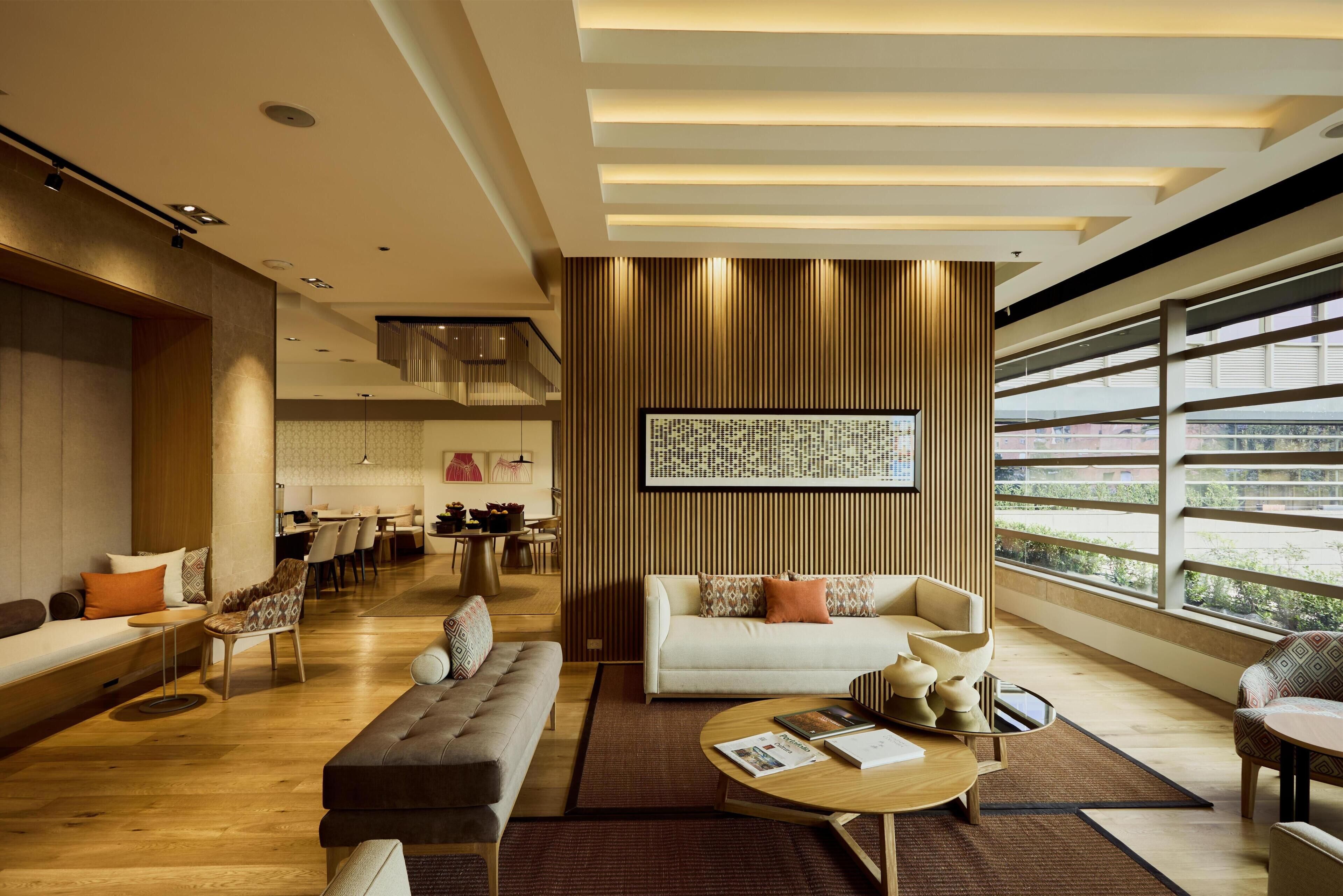 A modern living room with a sofa, coffee table, and chairs, a dining area in the background, and large windows with a view.