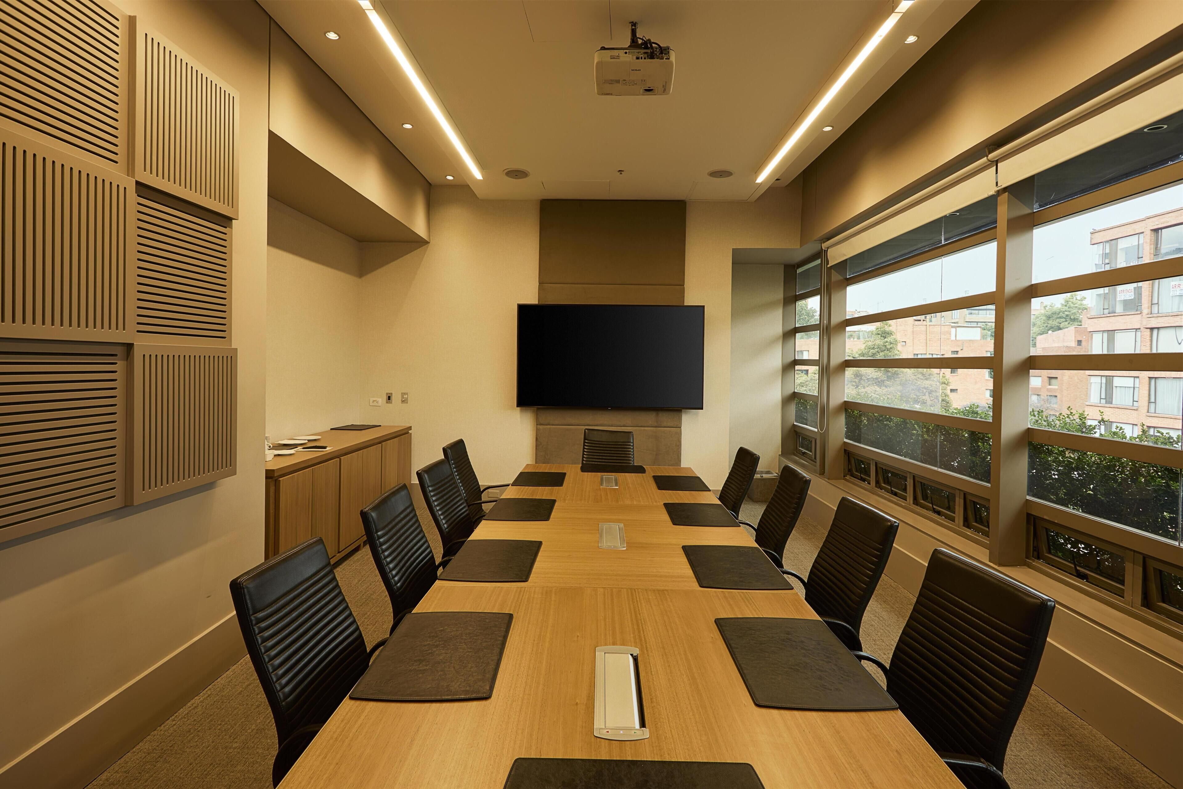 Un salón de conferencias con una larga mesa, sillas, un proyector y un amplio ventanal con vistas a edificios.