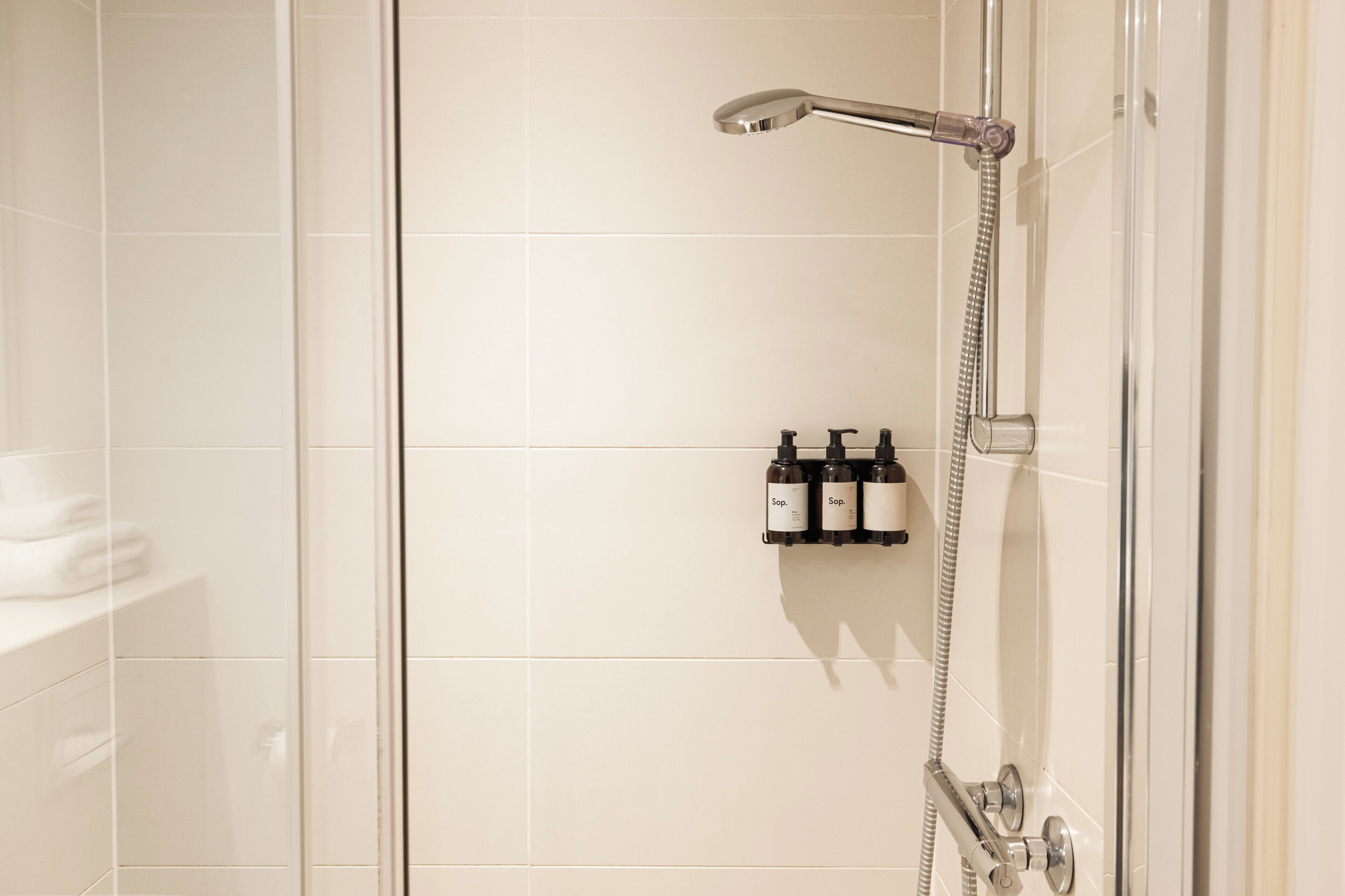 A shower with a handheld showerhead and a wall-mounted faucet. There are three bottles of soap on the wall.