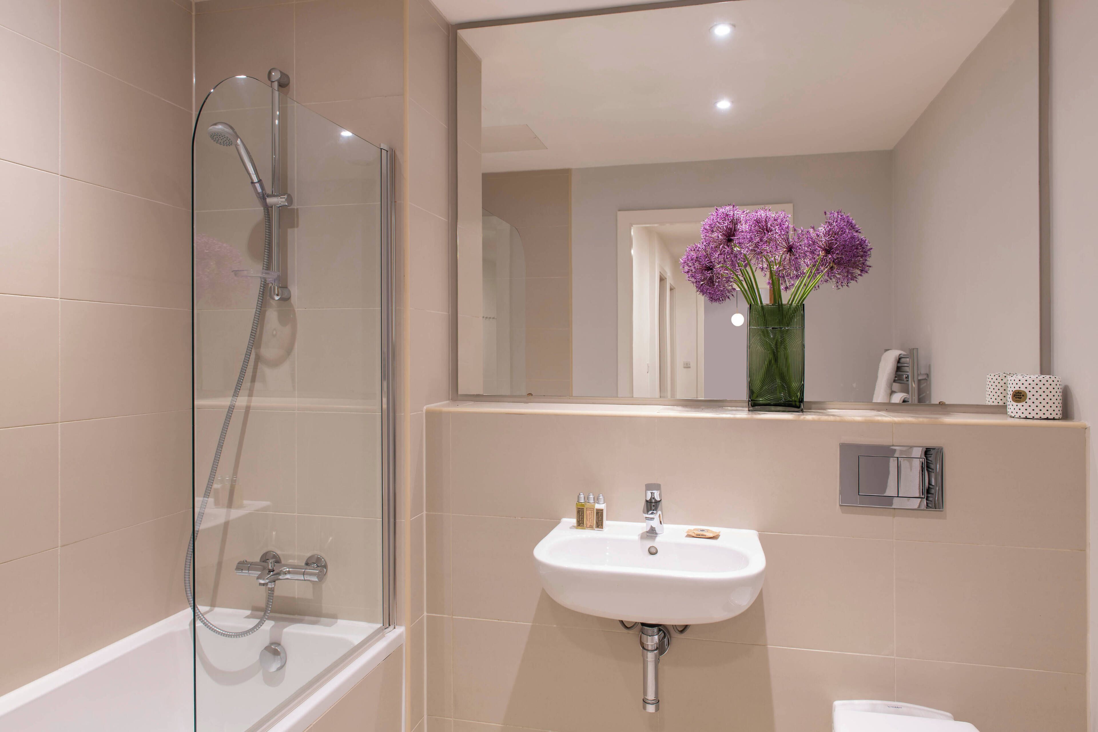 Un bagno moderno con doccia vetro, lavabo con specchio e un vaso con fiori viola.