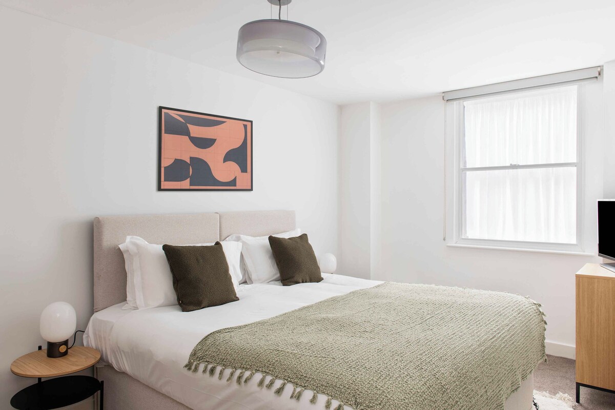 A neatly arranged bedroom with a bed, bedside table, lamp, artwork, and a window.