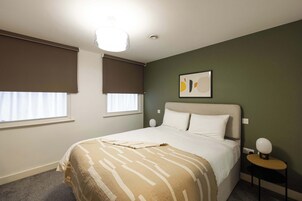 Un dormitorio con cama, dos ventanas con persianas, una mesita de noche con lámpara y una obra de arte colgada en la pared.
