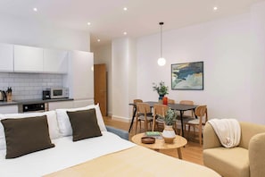 A modern hotel room with a bed, a dining table with chairs, a sofa, a kitchen area, and a painting on the wall.