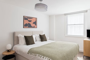 A bedroom with a bed, a bedside table, a lamp, a painting on the wall, and a window.