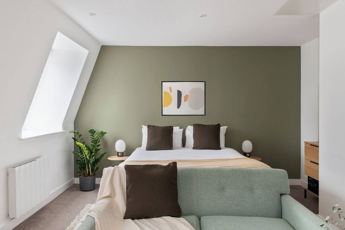 Ein modernes Schlafzimmer mit schrägem Dach, einem großen Bett mit vielen Kissen, einem Nachttisch mit Lampe, einer Topfpflanze und Blick ins Freie.