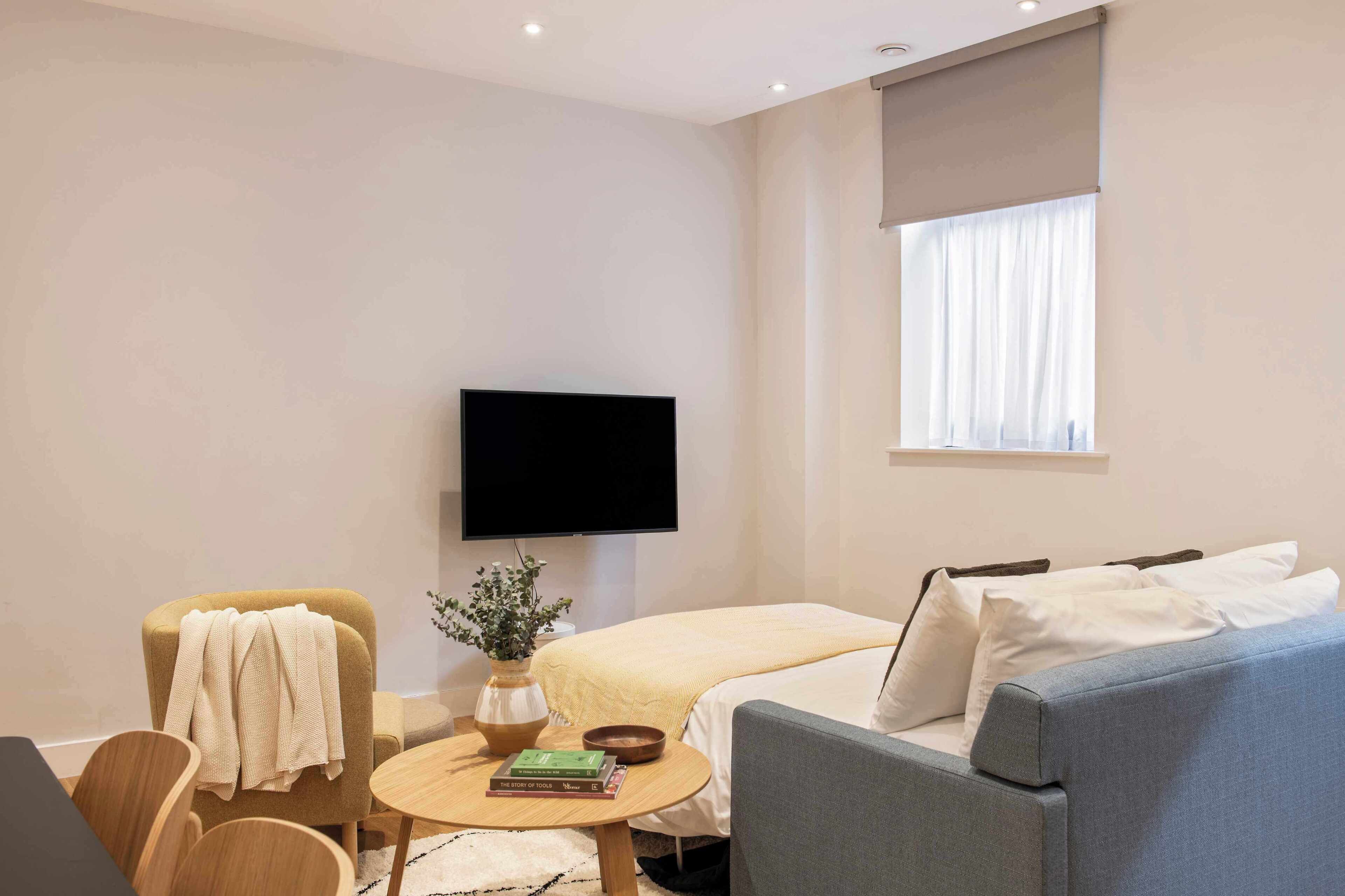 Une chambre d’hôtel moderne équipée d’un téléviseur à écran plat, d’un canapé, d’un fauteuil, d’une petite table ronde avec des livres et d’une fenêtre avec des rideaux.