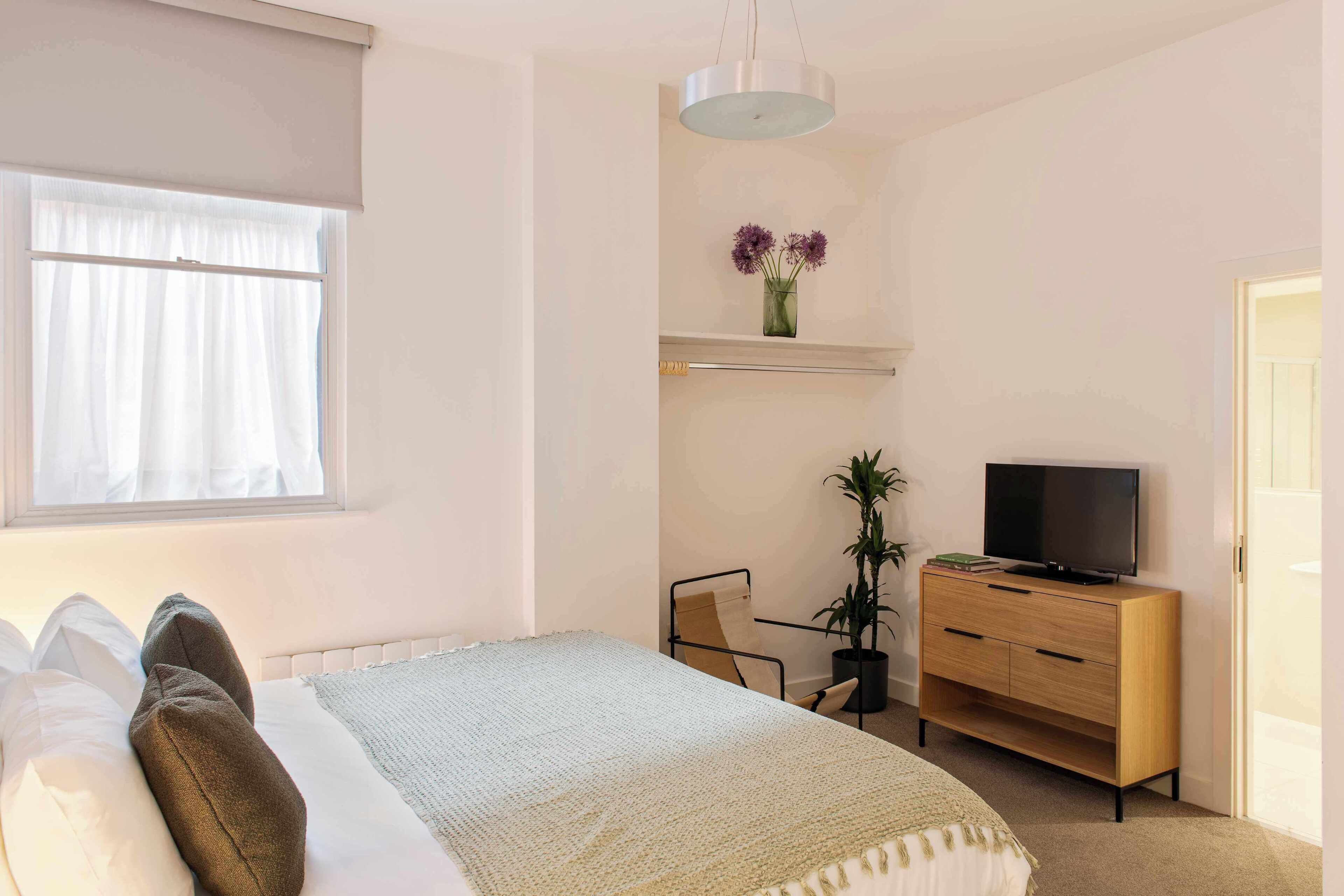 A bedroom with a bed, a TV on a wooden dresser, a chair, and a potted plant.