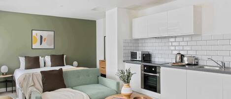 A modern apartment with a bed, a sofa, a small dining table, a kitchen with white cabinets and a microwave, and a green wall.