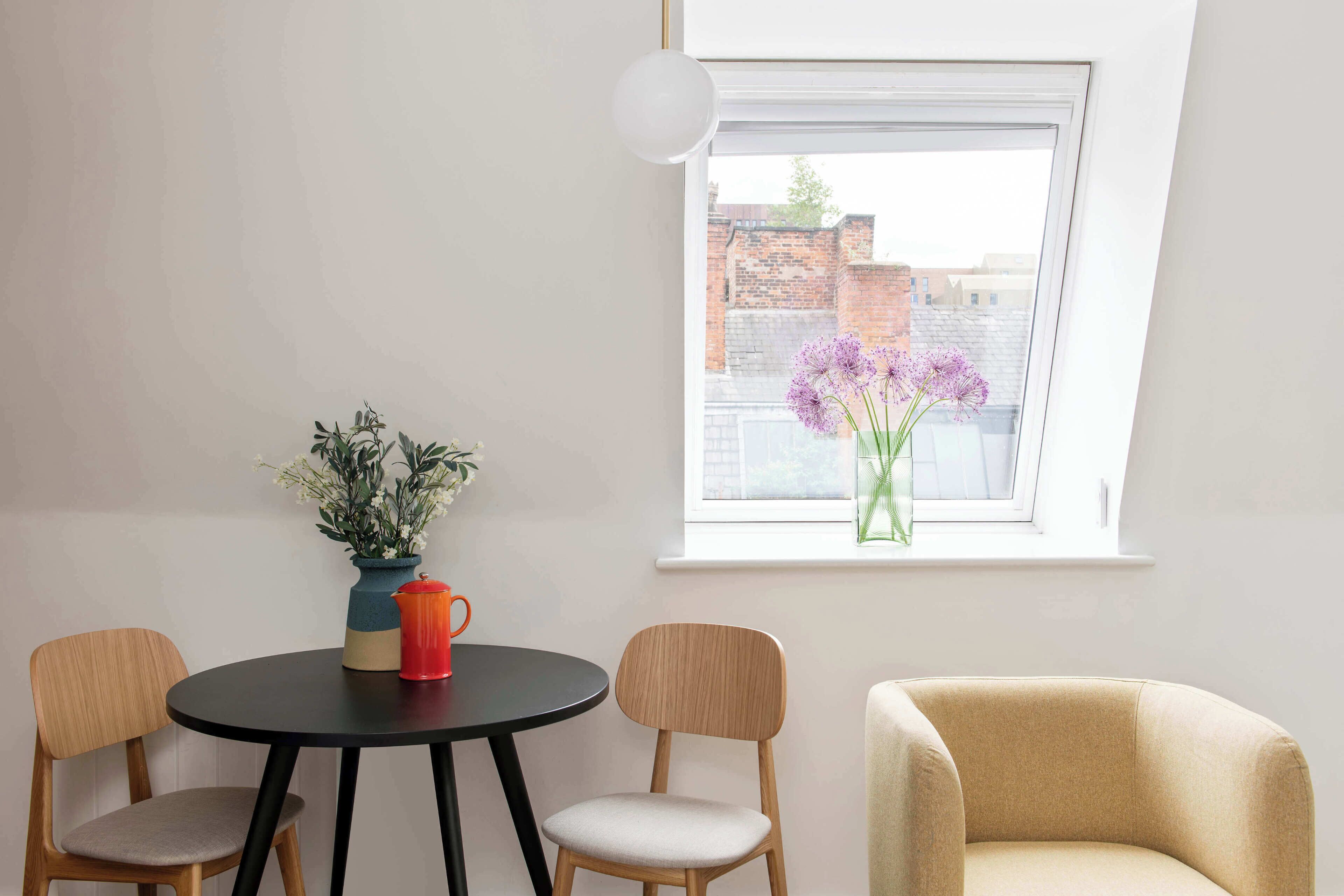 Ein Zimmer mit einem runden Tisch, zwei Stühlen, einer Vase mit Blumen und einem Fenster mit Blick auf Gebäude.
