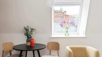 Ein Zimmer mit einem runden Tisch, zwei Stühlen, einer Vase mit Blumen und einem Fenster mit Blick auf Gebäude.