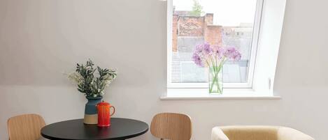 A room with a round table, two chairs, a vase with flowers, and a window with a view of buildings.