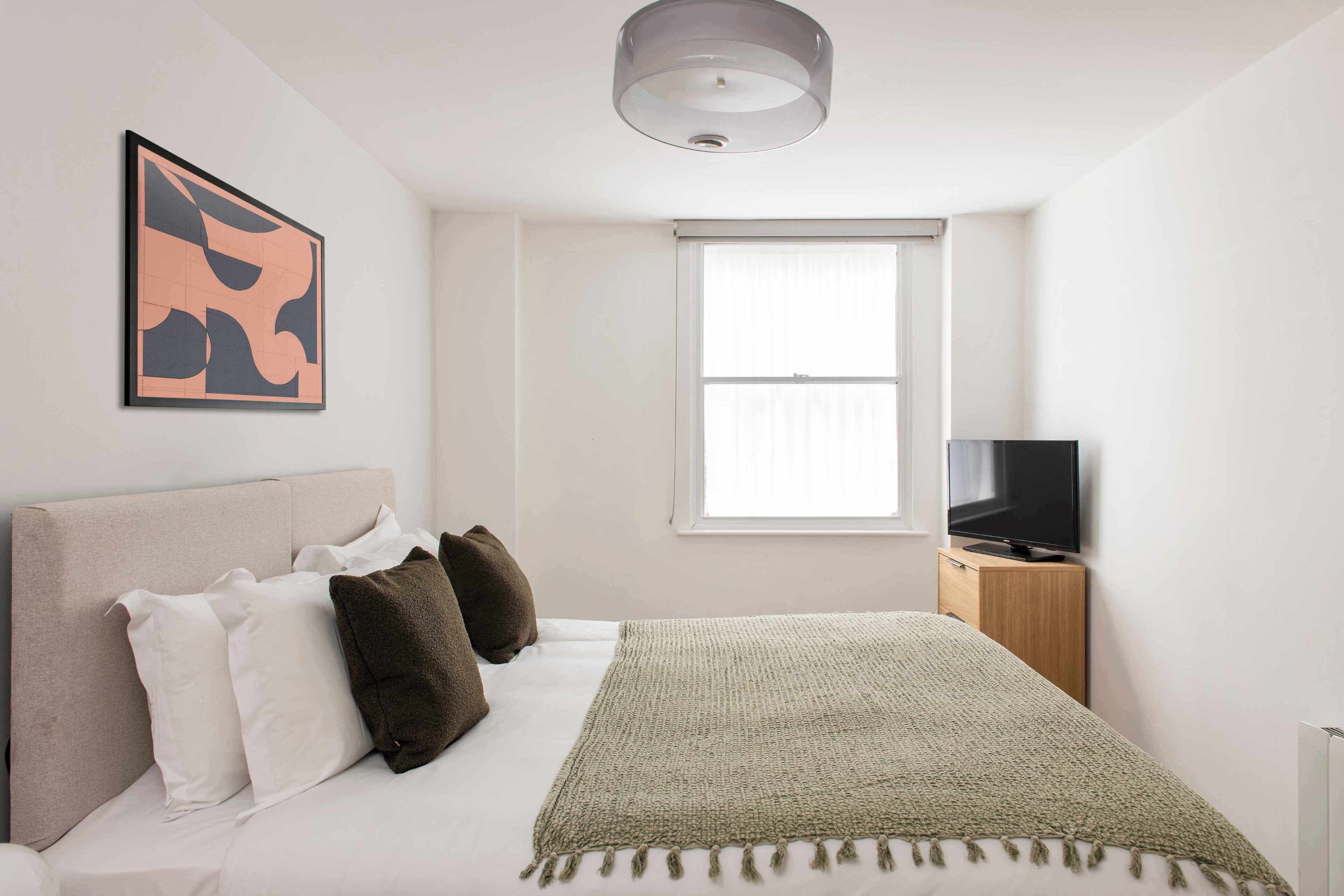 A bedroom with a bed, pillows, a blanket, a television on a wooden stand, a window, and a ceiling light.
