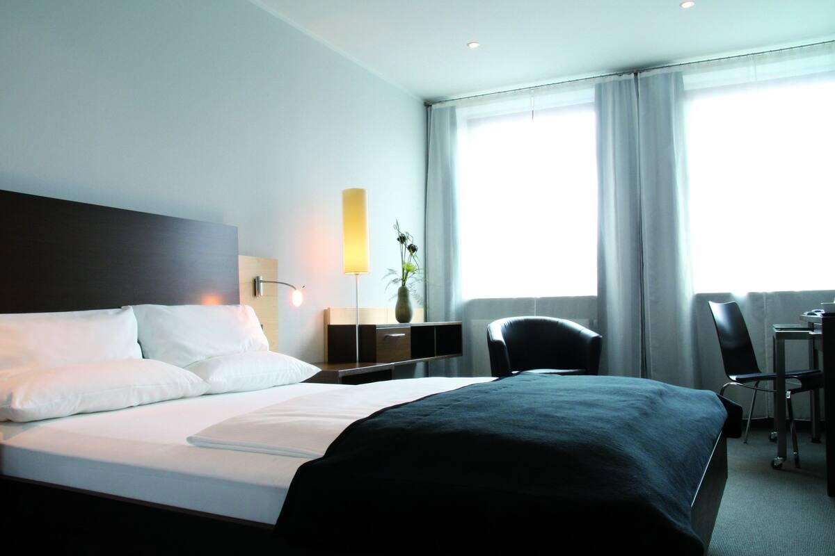A hotel room with a large bed, a desk, a chair, a lamp, and a window with curtains.